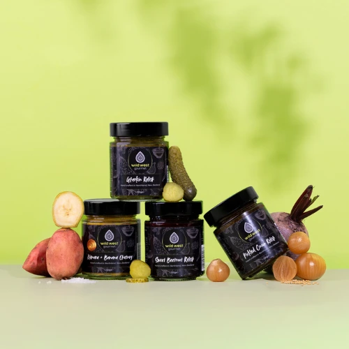 A line up of three relishes, with one leading jauntily on the jar to its left, and a fourth jar stacked on top. There is a crisp green backdrop.